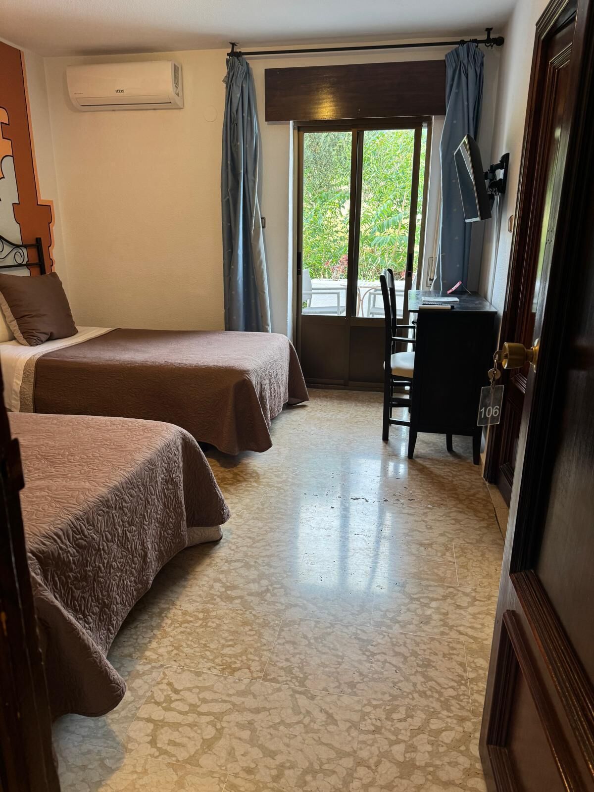 A hotel room with two beds, a desk, a chair, a TV, and a sliding glass door leading to an outdoor area.