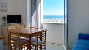 Una habitación con una mesa y sillas de madera, un televisor sobre un soporte y un ventanal con vistas a la playa y al océano.