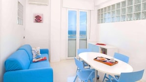 Una habitación luminosa con un sofá azul, una mesa blanca con sillas y vistas a través de un ventanal.