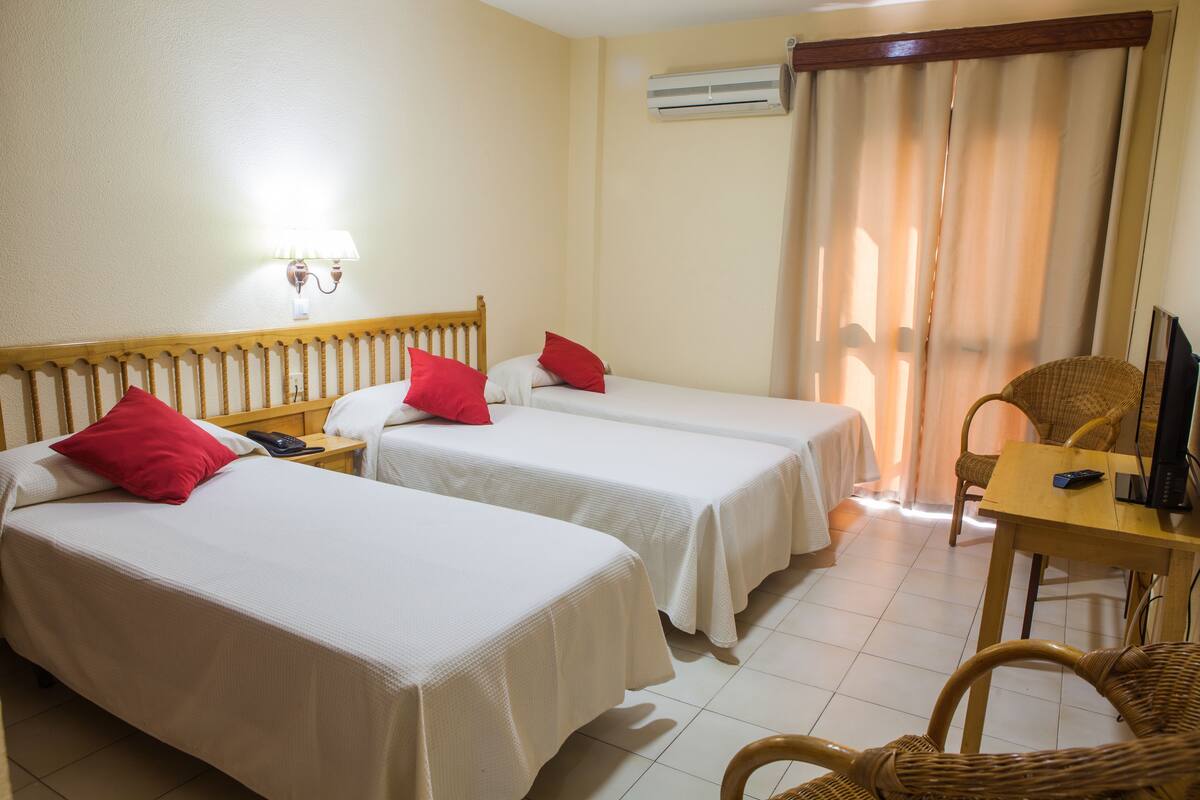 A hotel room with two single beds, a wooden headboard, a desk with a chair, and a window with curtains.