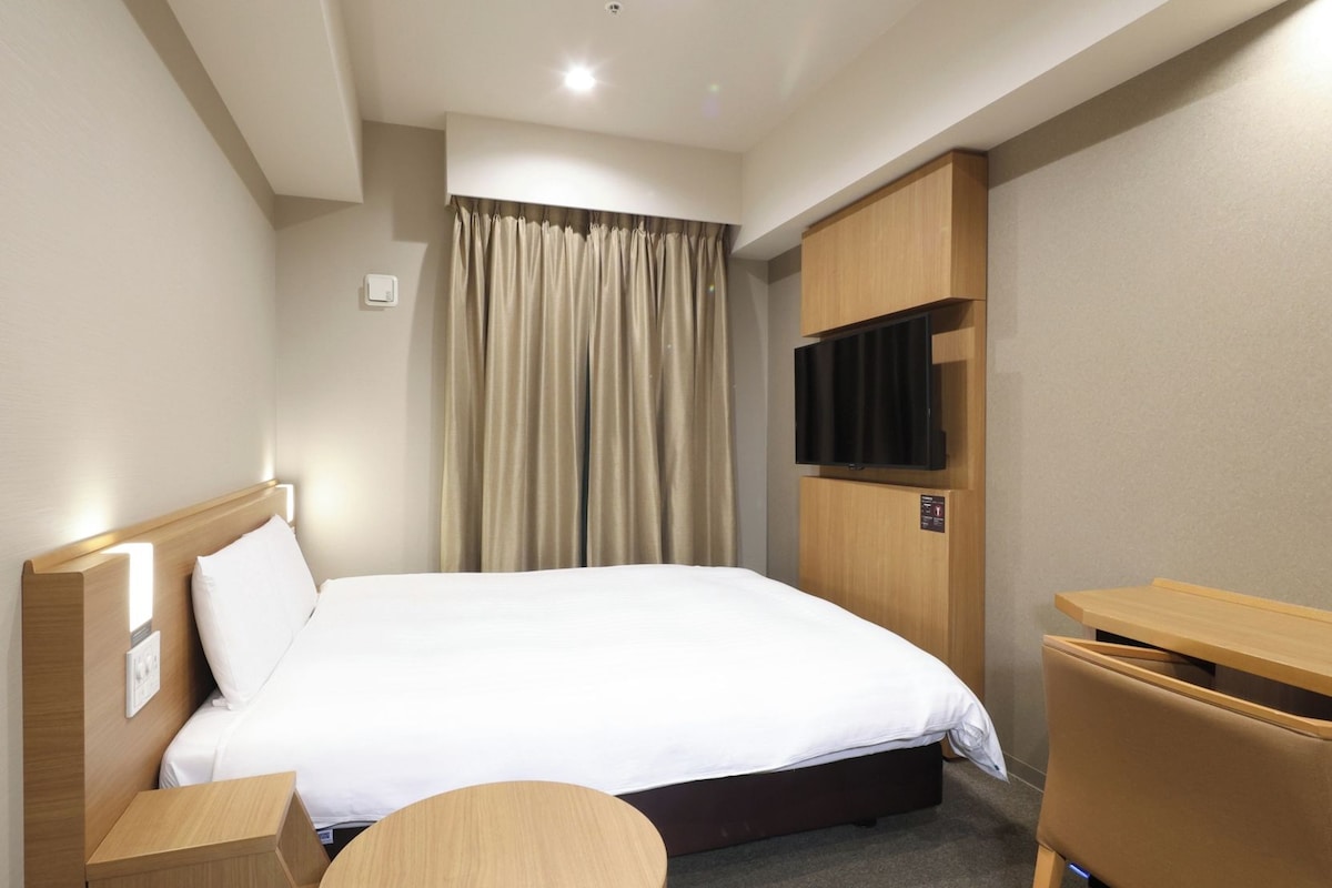 A hotel room with a bed, a desk, a chair, a round table, a television, and a curtain.