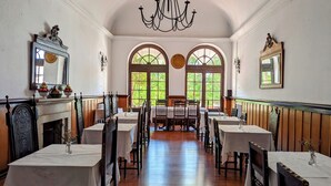 A dining room with wooden tables and chairs, a chandelier, and arched windows with greenery outside. - Hotel Solar De Monfalim (Évora)