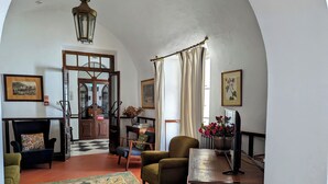 A cozy living room with a checkered floor, a large window with curtains, a wooden table with a vase of flowers, and a chair with a cushion. - Hotel Solar De Monfalim (Évora)