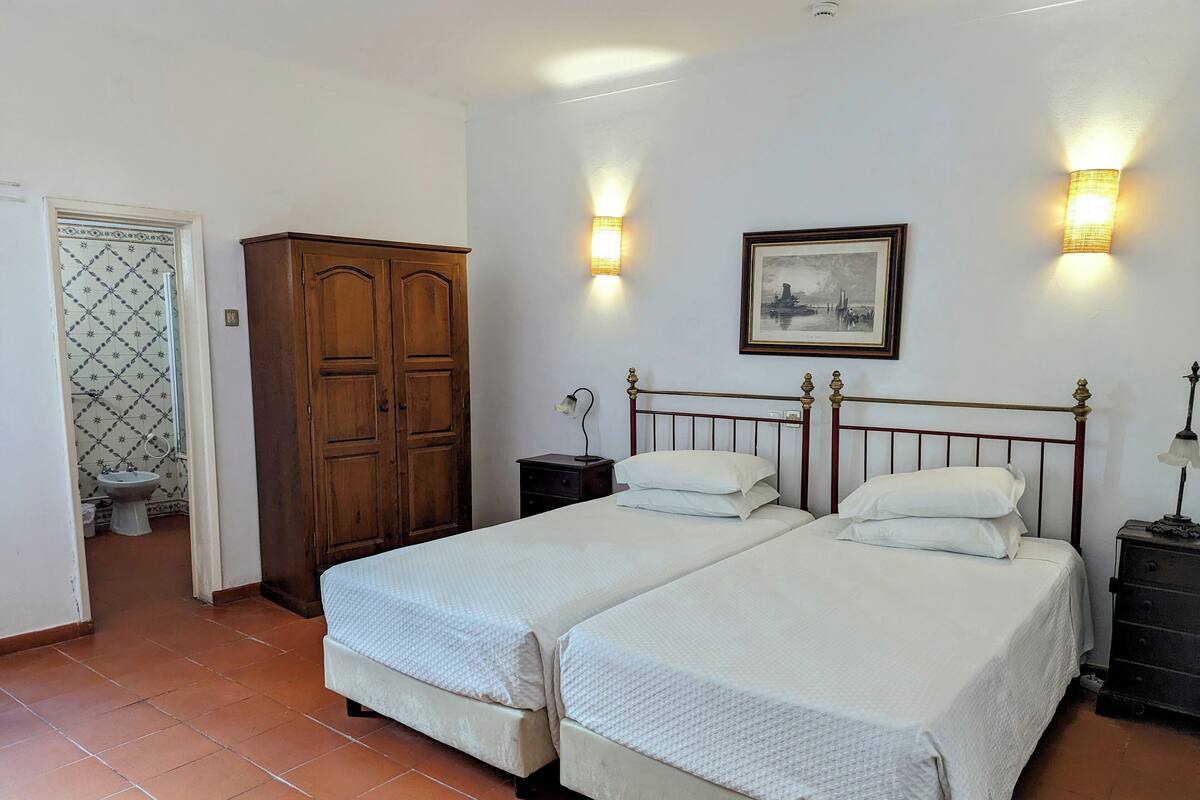 A bedroom with two beds, a wooden wardrobe, a bathroom with a tiled wall, and wall-mounted lights.