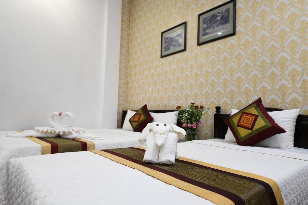 A hotel room with two beds, each with a white bedspread and a decorative pillow. There is a wall with a patterned wallpaper and two framed pictures.