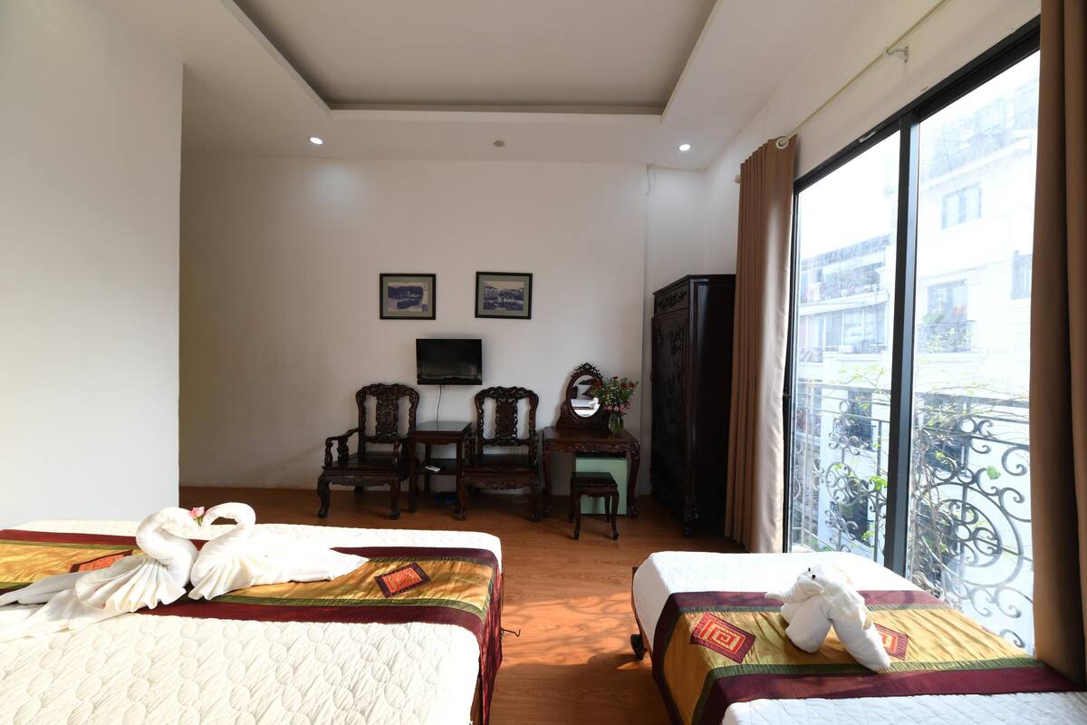 A hotel room with two beds, a desk with chairs, a television, and a balcony with a view of buildings.