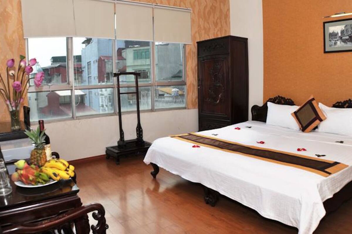 A hotel room with a large bed, a wooden desk with a fruit bowl, a standing lamp, and a window with a city view.