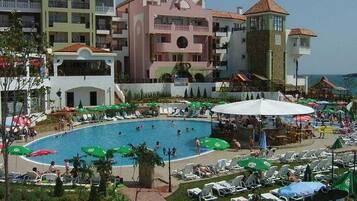 A hotel with a swimming pool surrounded by lounge chairs and umbrellas, a landscaped garden, and multi-story buildings with balconies.