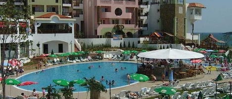 A hotel with a swimming pool surrounded by lounge chairs and umbrellas, a landscaped garden, and multi-story buildings with balconies.