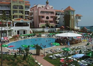 A hotel with a swimming pool surrounded by lounge chairs and umbrellas, a landscaped garden, and multi-story buildings with balconies. - Bella Vista Beach Club (Tsarevo)