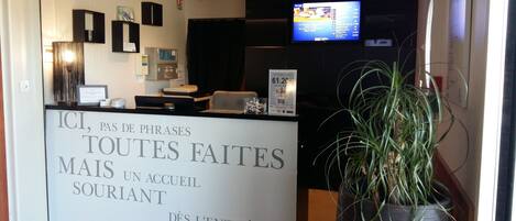 A hotel reception desk with a sign in French, a television screen, and a potted plant.