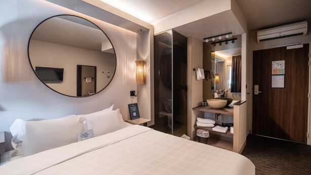 Habitación de hotel con una cama grande, un espejo redondo, televisor integrado, baño con lavamanos y espejo, y un equipo de aire acondicionado.