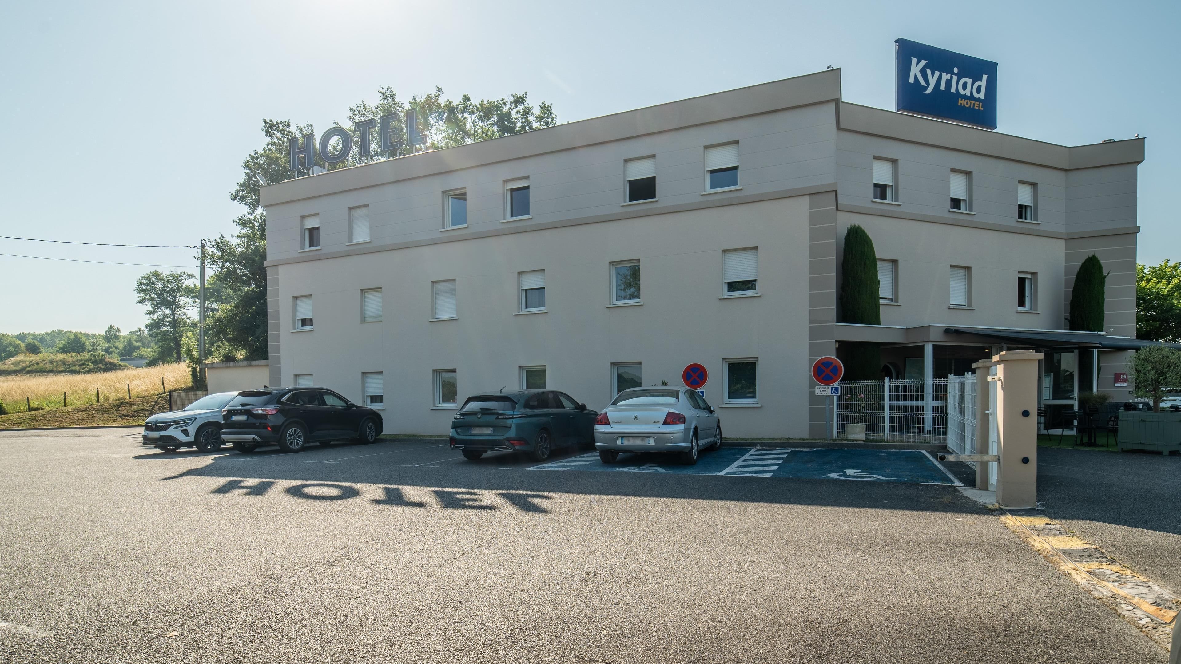 Un hotel llamado 'Kyriad Hotel' con un edificio moderno, una zona de estacionamiento y una entrada protegida por una puerta.