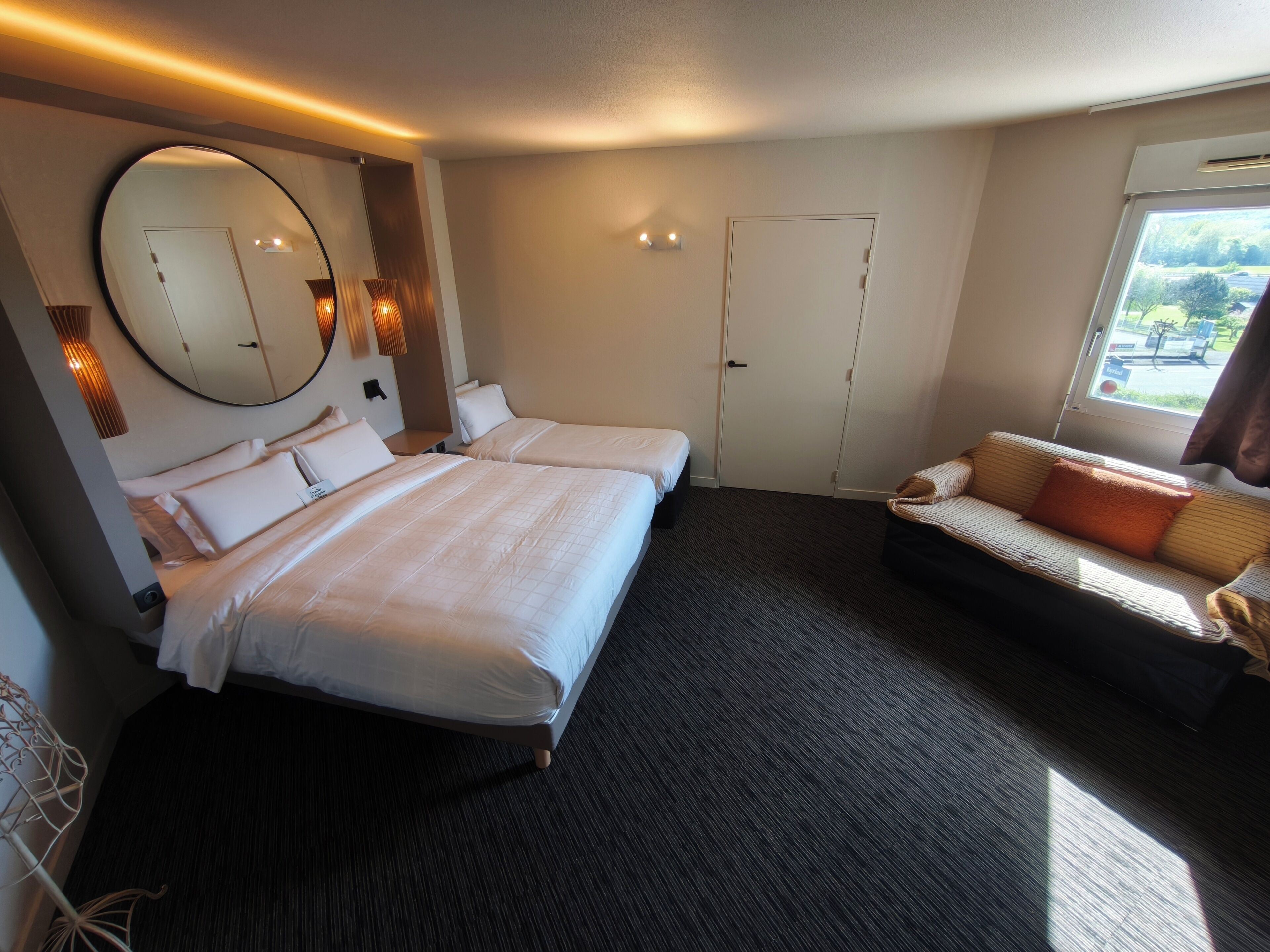 A hotel room with two beds, a sofa, a large mirror, and a window with a view.