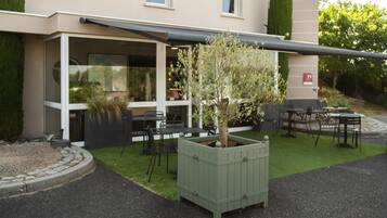 Ein Außenbereich mit Tischen und Stühlen, ein grüner Pflanzkübel mit einem Baum und ein Gebäude mit großen Fenstern und einer überdachten Terrasse.