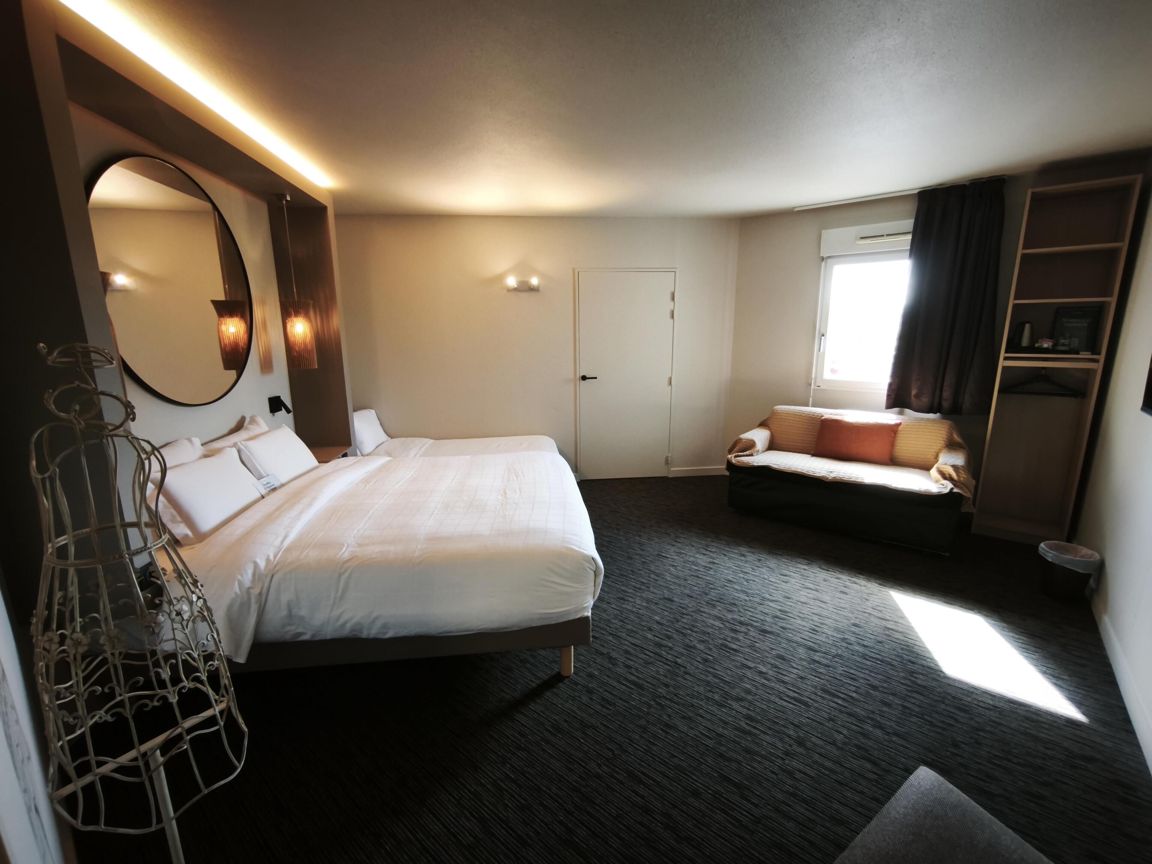 A hotel room with a large bed, a sofa, a mirror, a wardrobe, and a window with curtains.