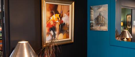 A room with a framed abstract painting, a lamp, and a mirror reflecting a building.