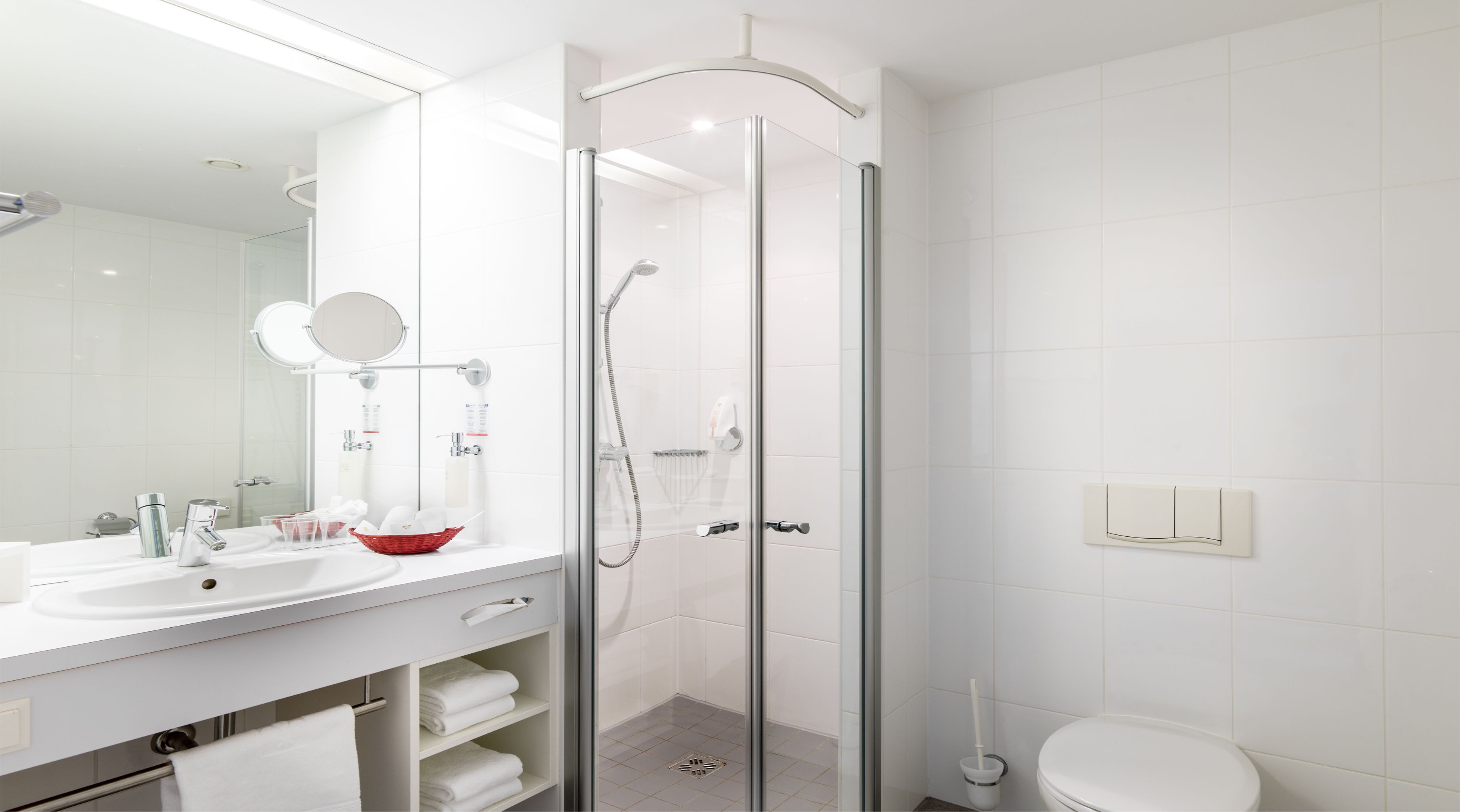 Un baño moderno con ducha envuelta en cristal, un espejo grande, un lavamanos con un jabonera roja y un inodoro.