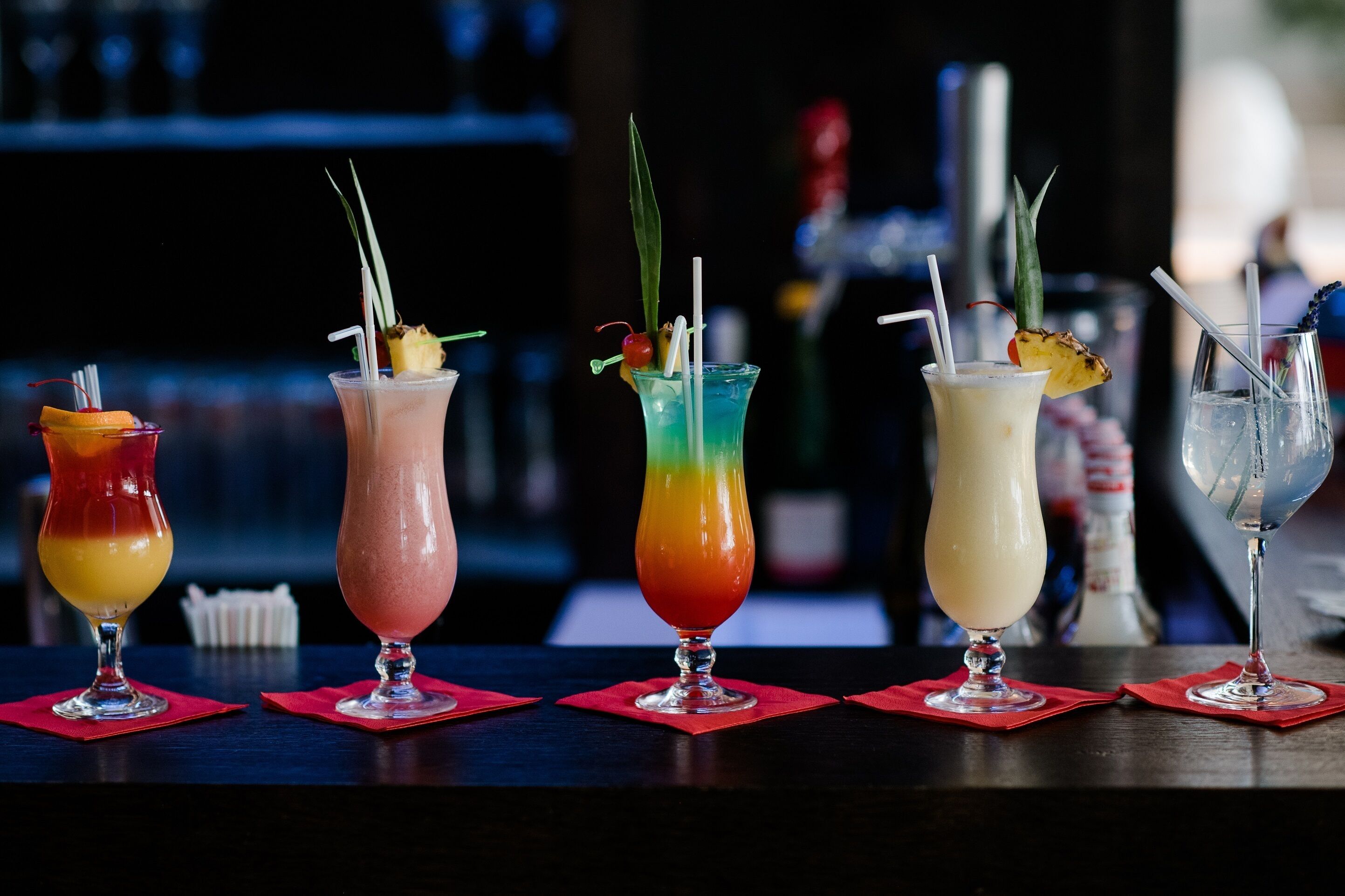 A bar with five colorful cocktails, each garnished with a straw and a decorative fruit.