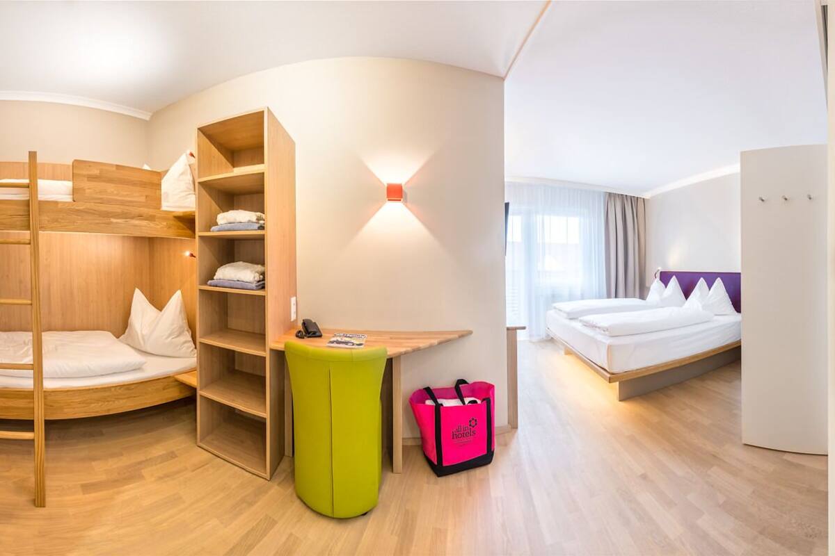 A hotel room with bunk beds, a desk, a green ottoman, a suitcase, and a bed with a purple headboard.