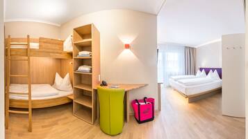 Habitación de hotel con literas, un escritorio, un reposapiés verde, una maleta y una cama con cabecera morada.