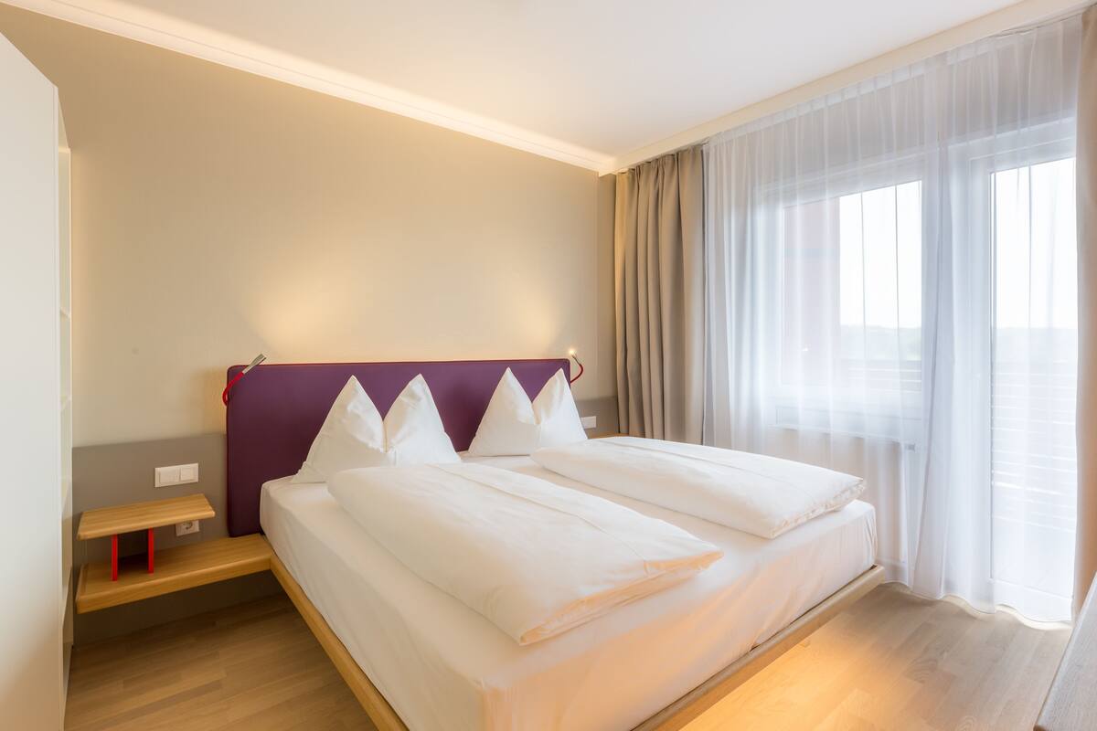 A modern hotel room with a large bed, a bedside table, and a window with sheer curtains.