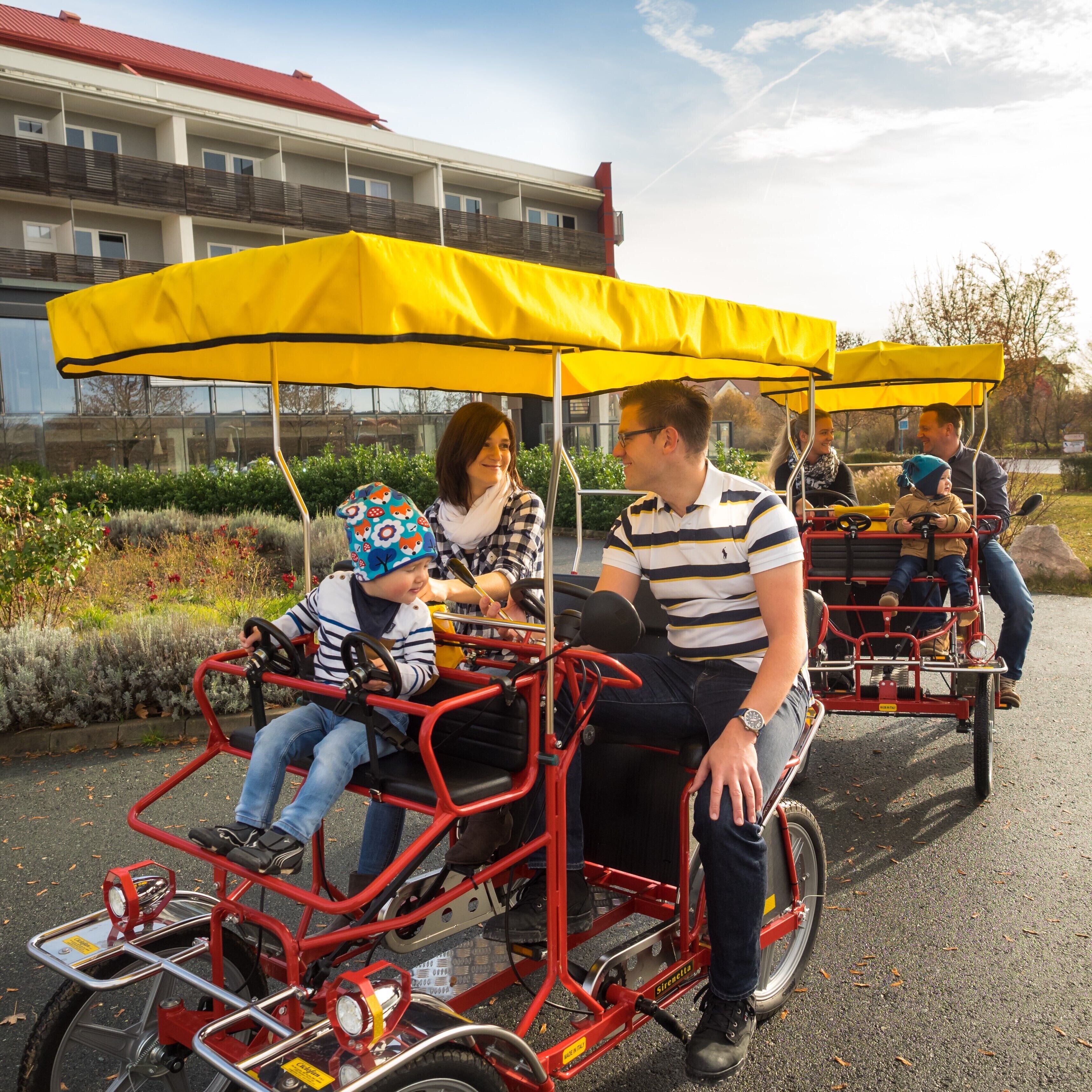 Een rode koets met een geel zonnedak, passagiers en een gebouw op de achtergrond.