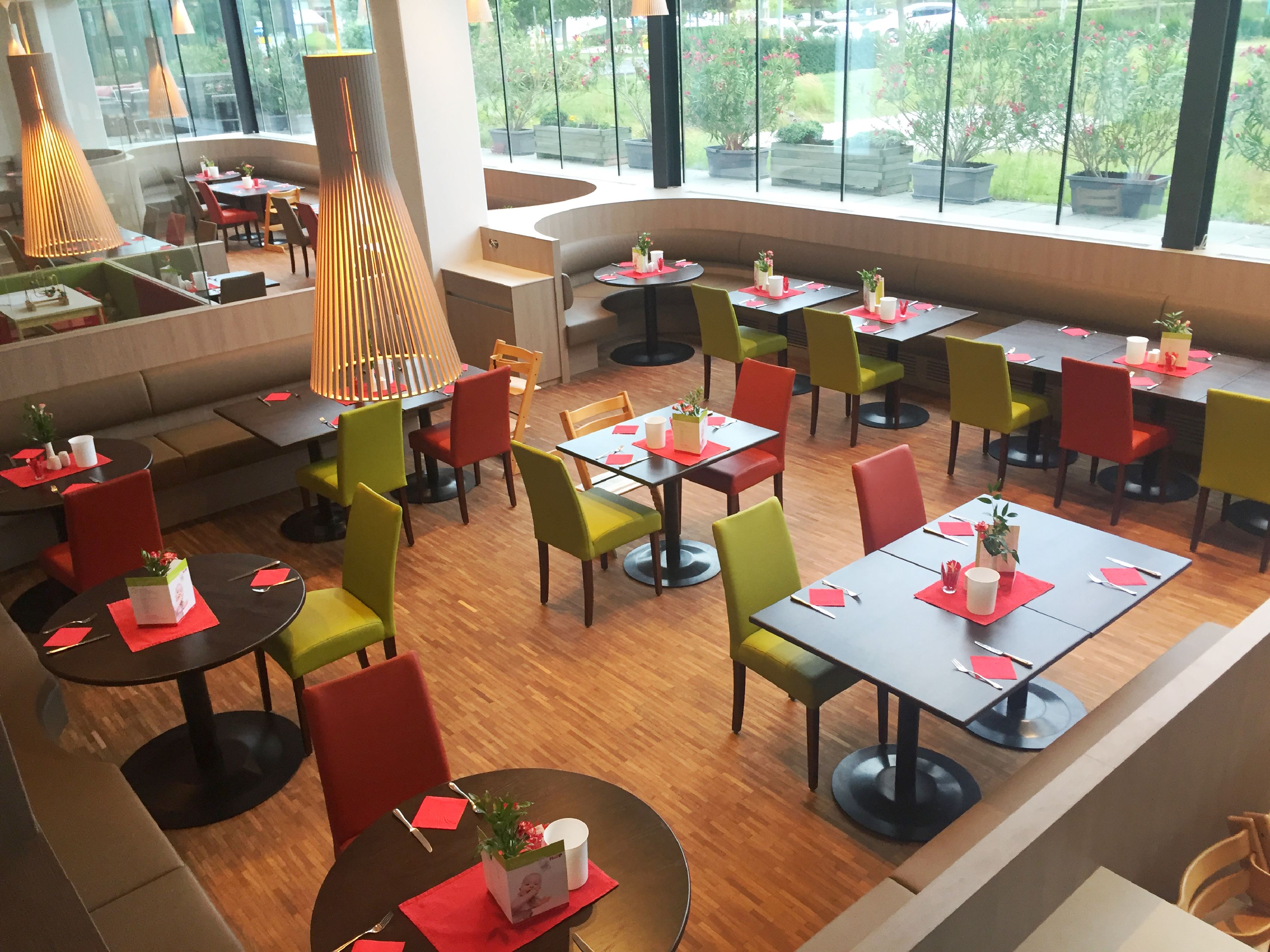 A modern restaurant with round tables and colorful chairs, large windows offering a view of the garden, and a central light fixture.