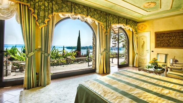 A luxurious bedroom with a large window overlooking a scenic view, a bed with a striped bedspread, and a classic chandelier. - Park Hotel Ville Montefiori (Gardone Riviera)