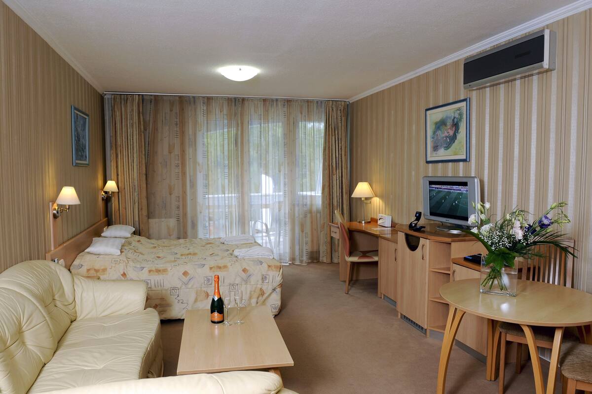 A hotel room with a bed, a sofa, a desk with a computer, a TV, and a small table with a bottle and glasses.