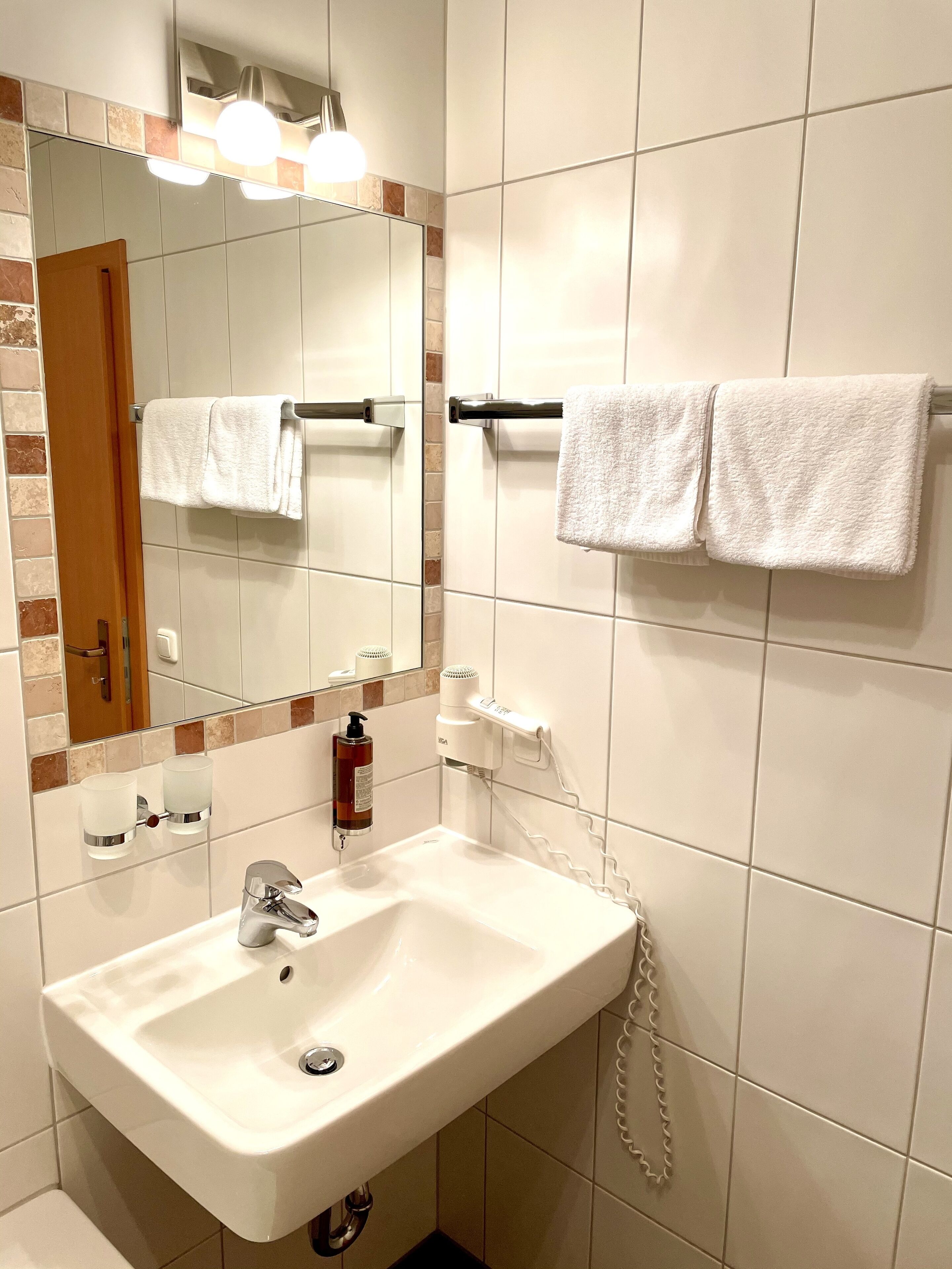 Baño con lavamanos blanco, espejo, toallero con dos toallas, secador de pelo y jabón de manos.