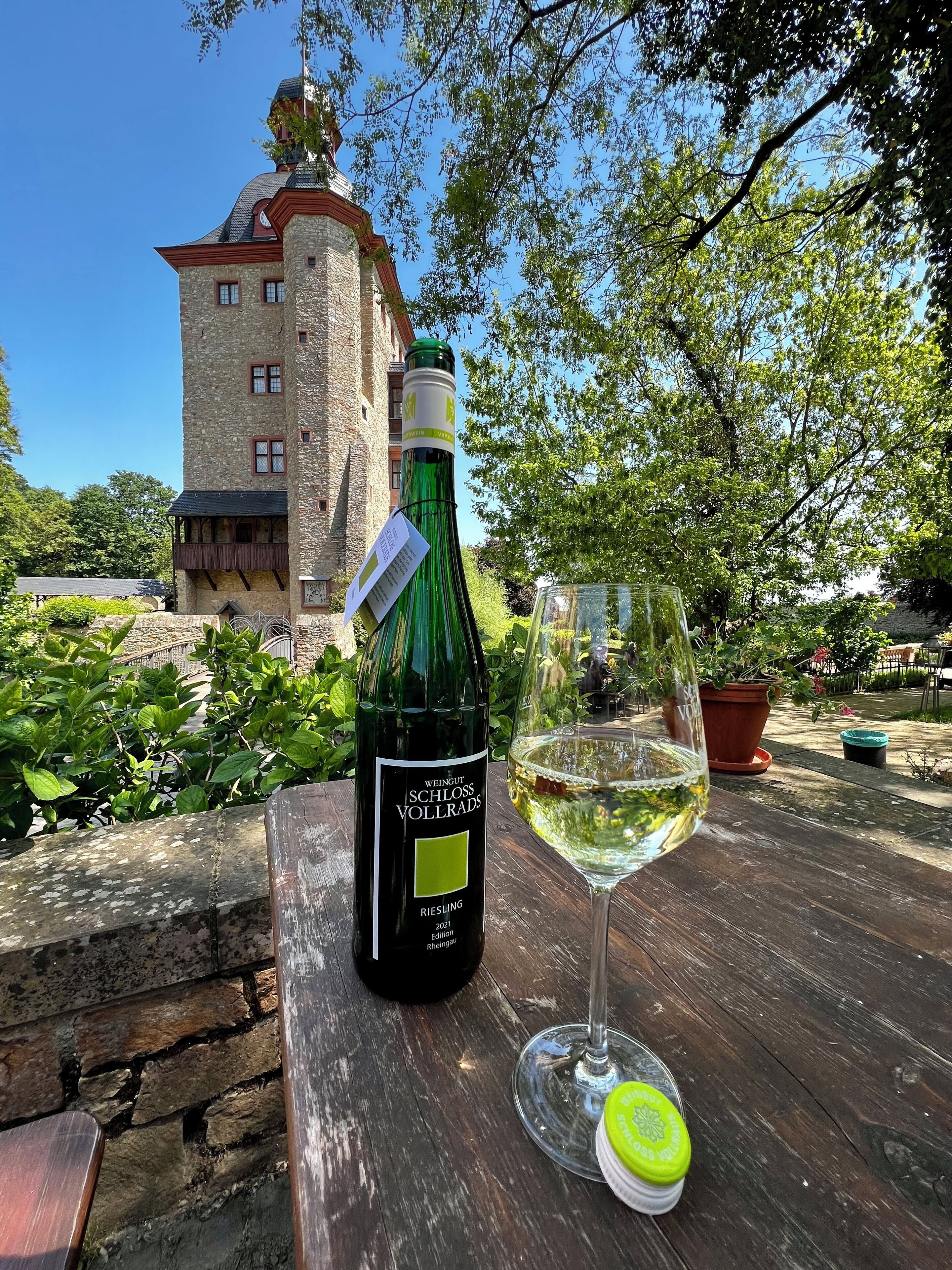 Una botella de vino Riesling y una copa sobre una mesa de madera, con un edificio de piedra al fondo.