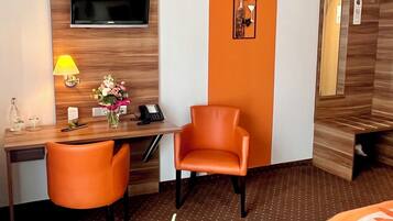 A hotel room with a wooden desk, a television mounted on the wall, an orange chair, a small table with a lamp, and a bed with a floral bedspread.