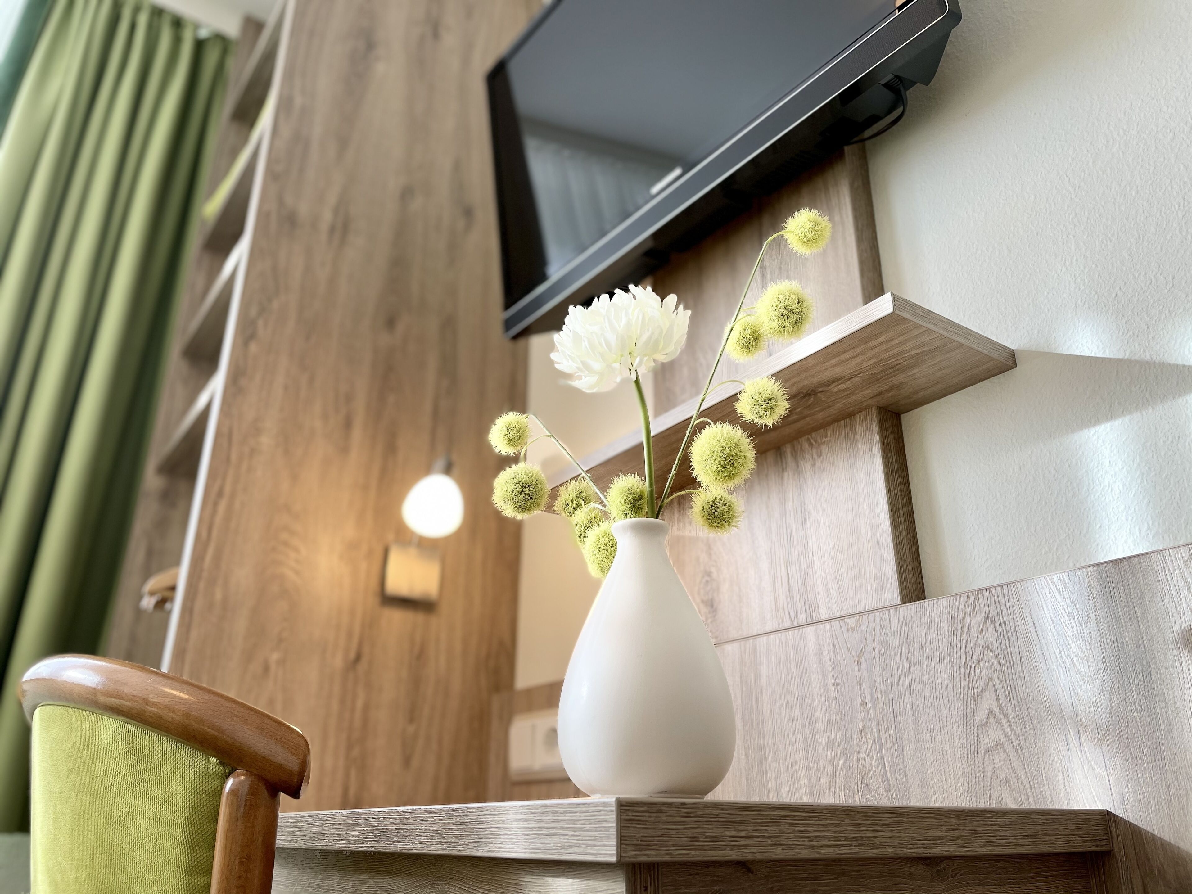 A white vase with flowers on a wooden shelf, a television mounted on the wall, and a green chair.