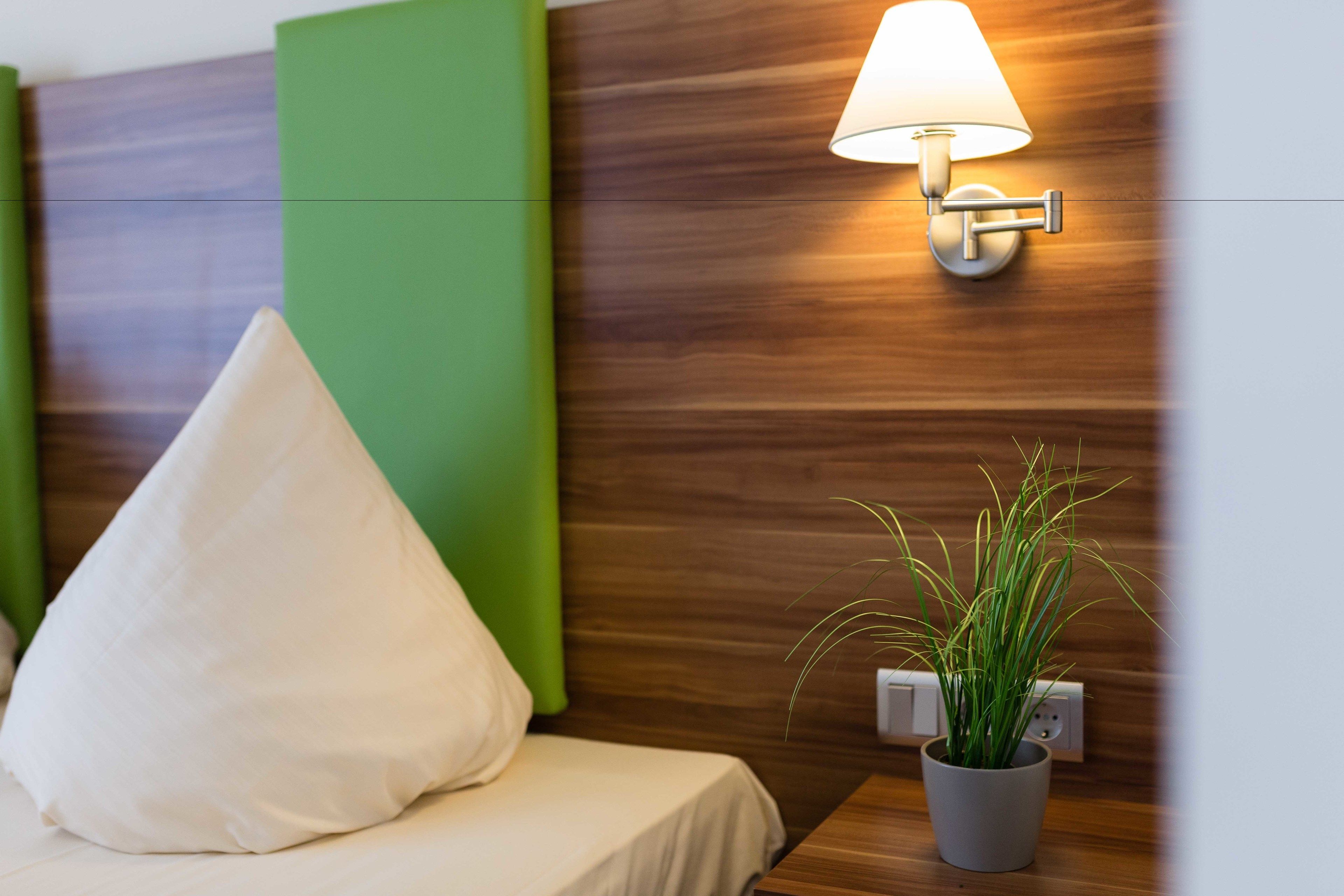 Quarto de hotel com uma cama, parede de destaque verde, luminária fixada na parede e planta em vaso.