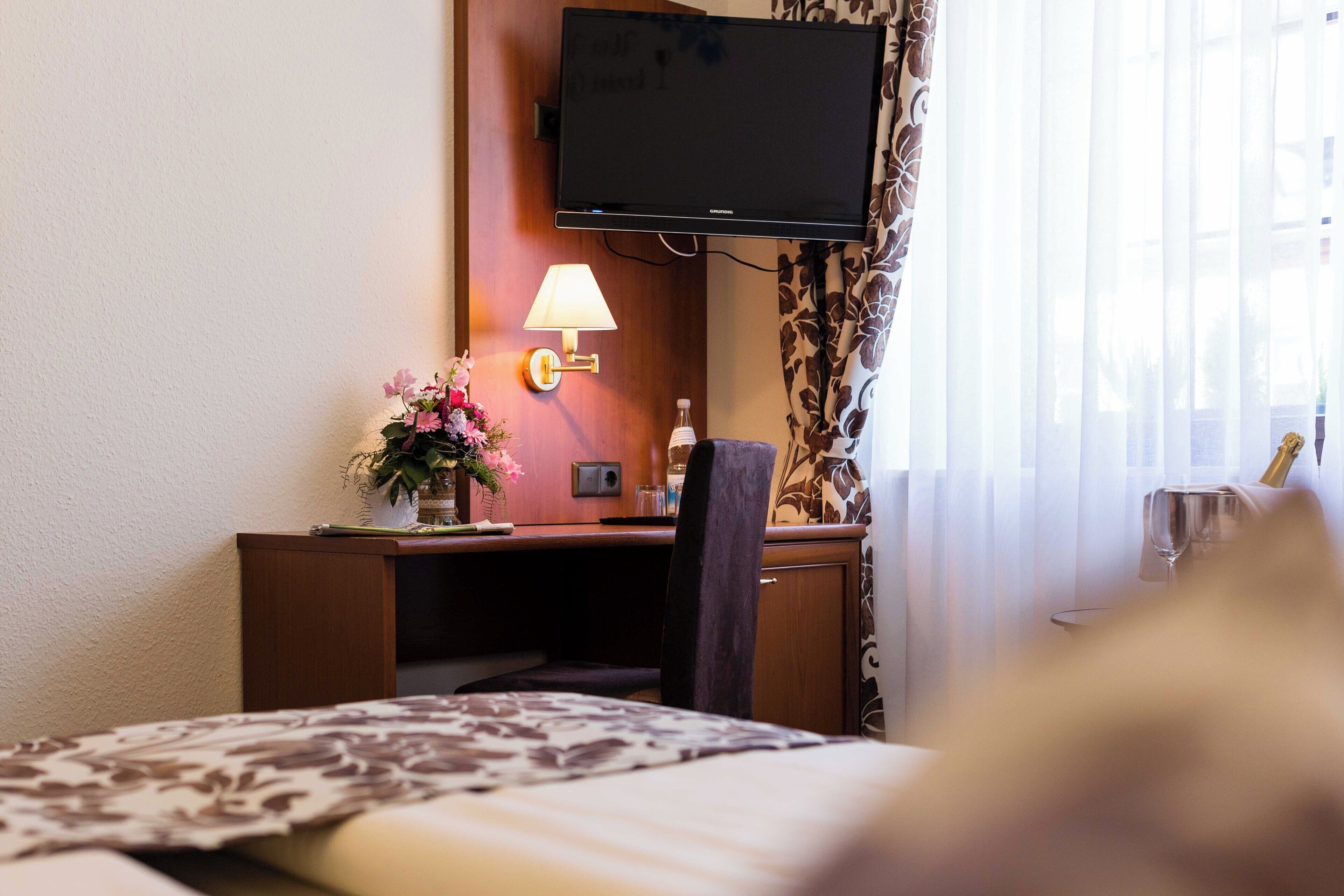 Une chambre d’hôtel avec un lit, un bureau avec une lampe, une télévision et une fenêtre avec des rideaux.