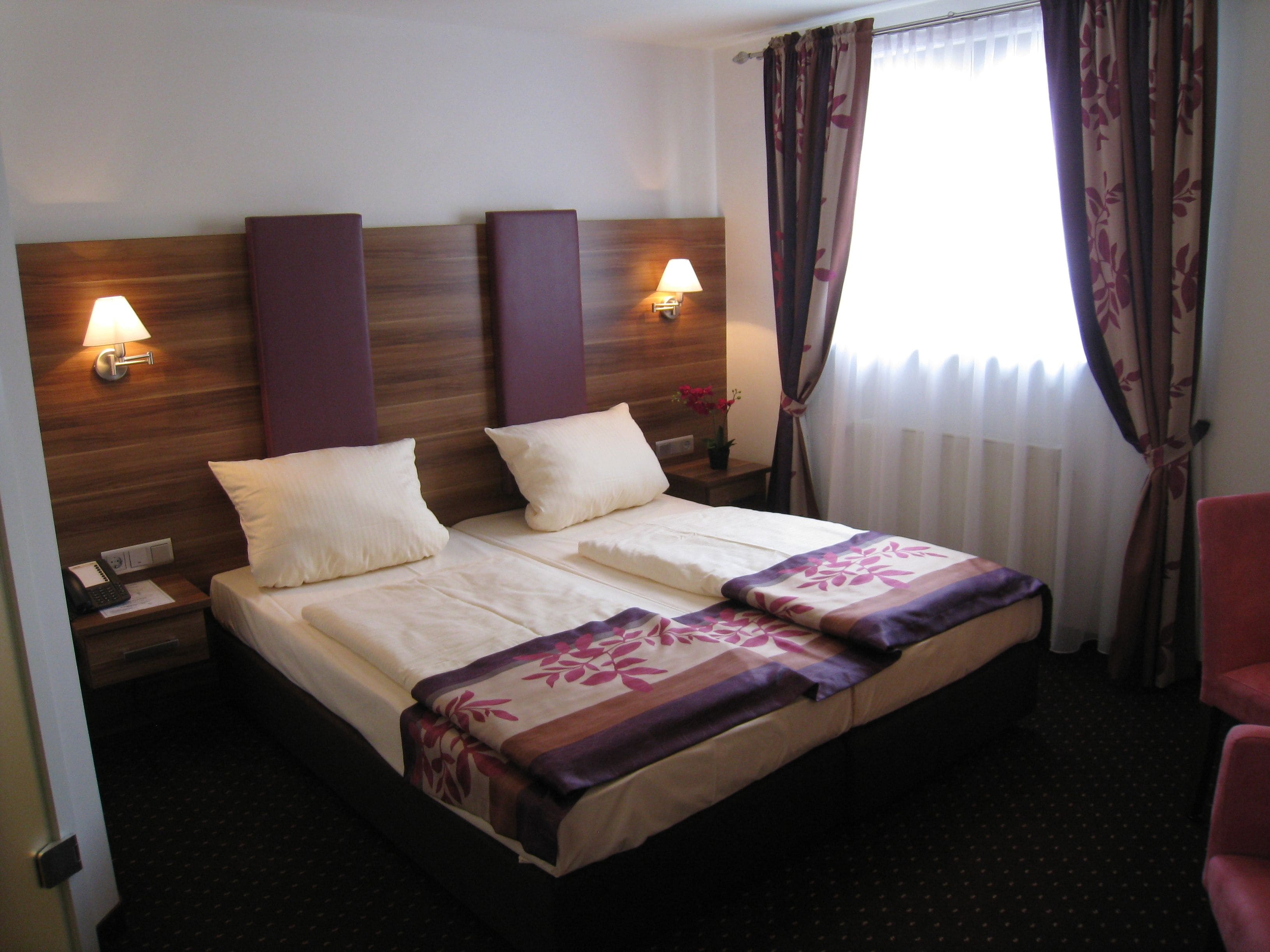 Quarto de hotel com cama, duas luminárias de cabeceira, mesa de apoio, telefone e janela com cortinas.