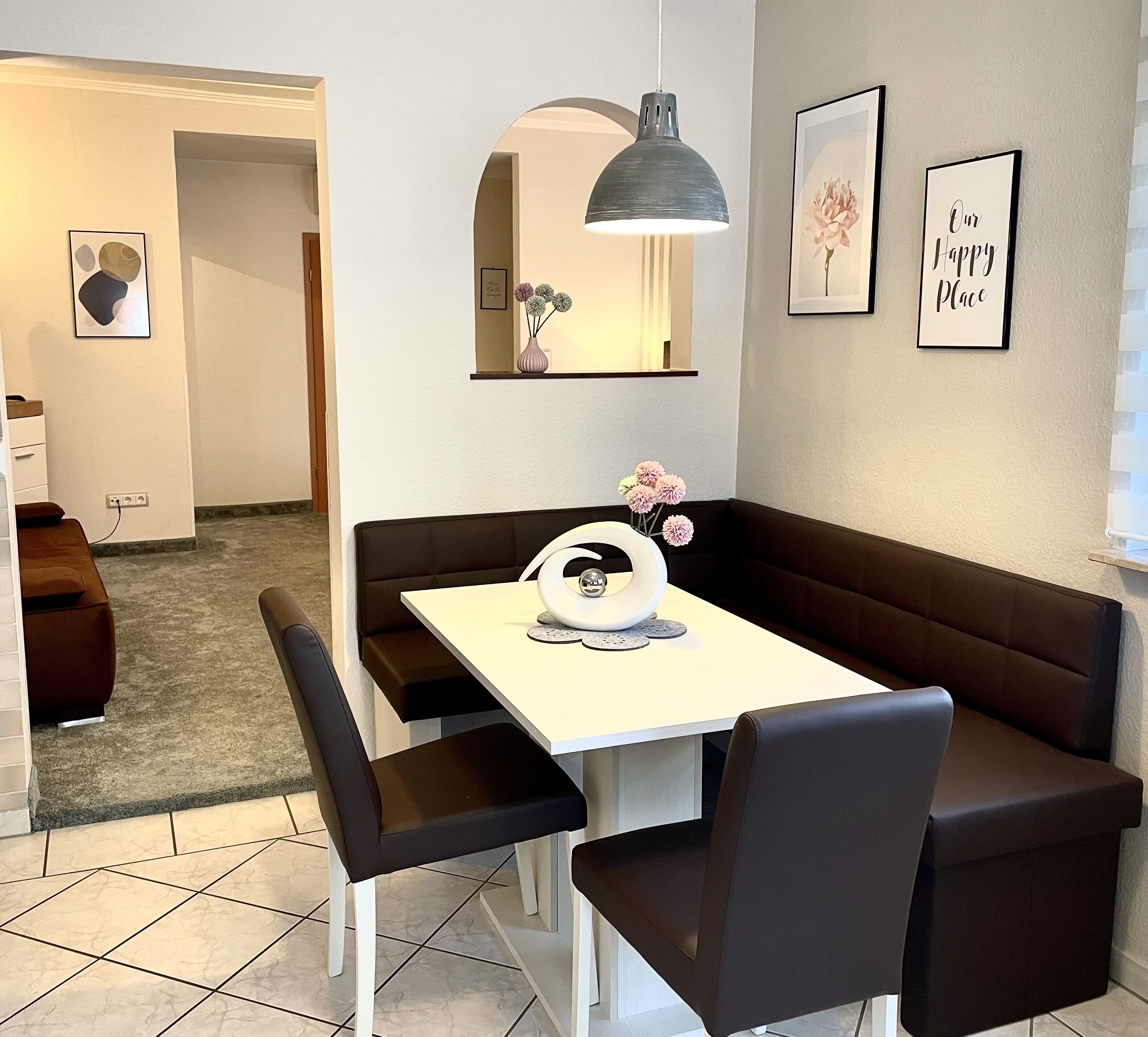 Un comedor moderno con una mesa blanca, sillas negras y una lámpara colgante oscura. Hay cuadros en las paredes y un banco a lo largo de la pared.