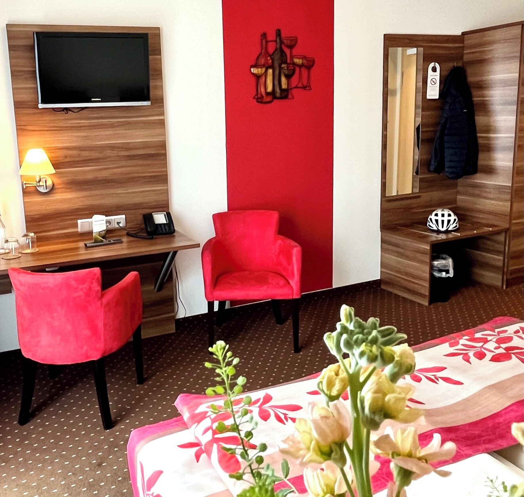 A hotel room with a bed, a desk with a chair, a TV, a red wall, and a coat rack.