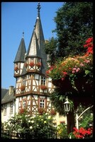 Um edifício tradicional em enxaimel com várias torres, rodeado por vegetação exuberante e plantas floridas.