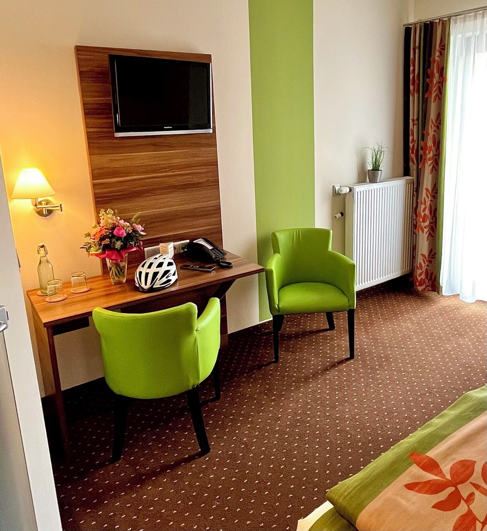 Habitación de hotel con escritorio de madera, dos sillas verdes, televisor en la pared, radiador y ventana con cortinas.