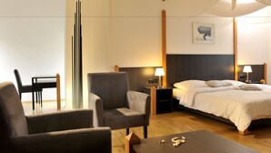 Habitación de hotel con una cama, dos mesitas de noche con lámparas, una silla, una pequeña mesa con adornos y una lámpara de techo alta y moderna.