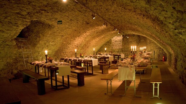 Un comedor subterráneo con paredes de piedra, mesas y bancos de madera, y mesas iluminadas con velas.