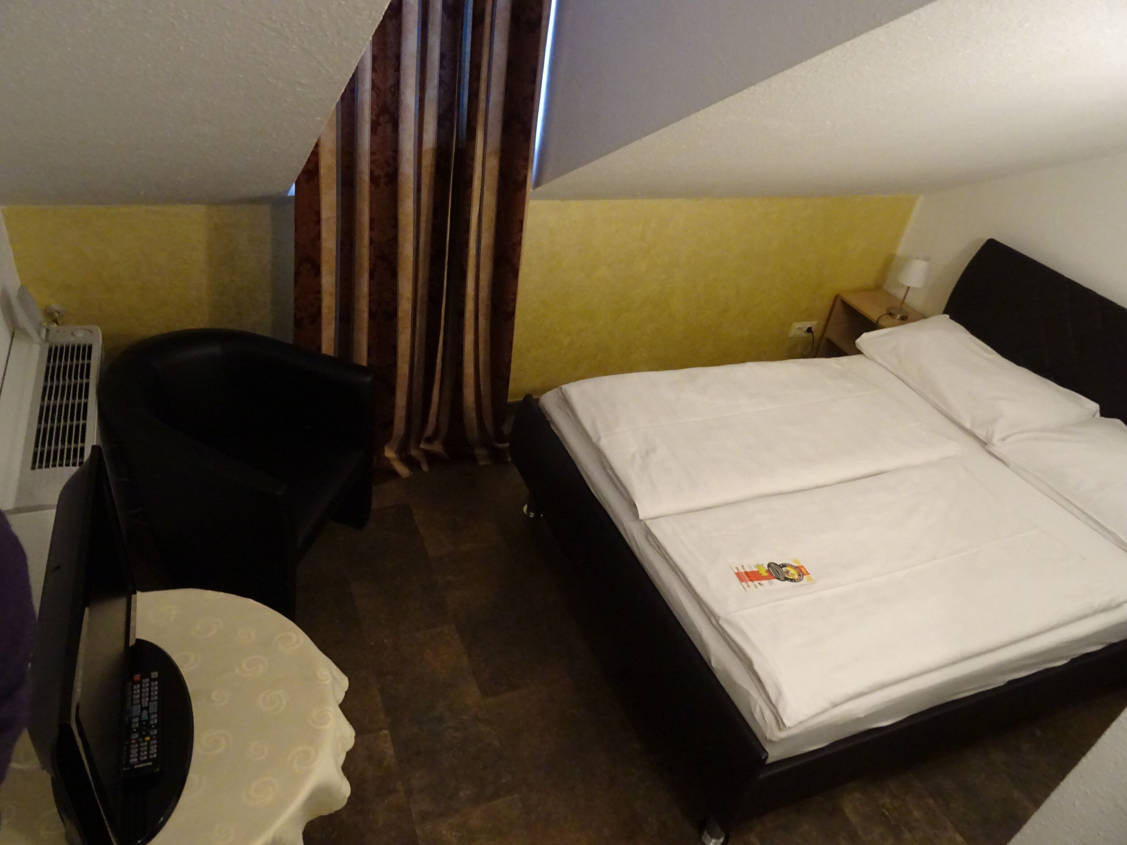 A hotel room with a bed, a chair, a small table, and a curtain.