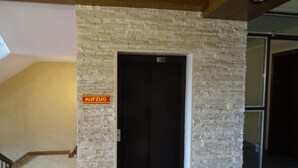 A room with a white brick wall, a door with a sign reading "AUFZUG", and a staircase leading to another area. - Apparthotel Alte Innbruecke (Neuhaus am Inn)