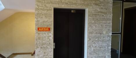 Una habitación con pared de ladrillo blanco, una puerta con un cartel que dice "AUFZUG" y una escalera que conduce a otra zona.