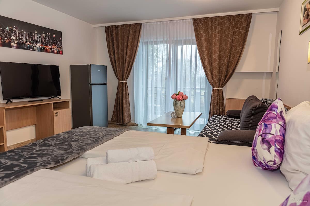 A hotel room with a bed, a desk with a TV, a refrigerator, a window with curtains, and a vase with flowers.