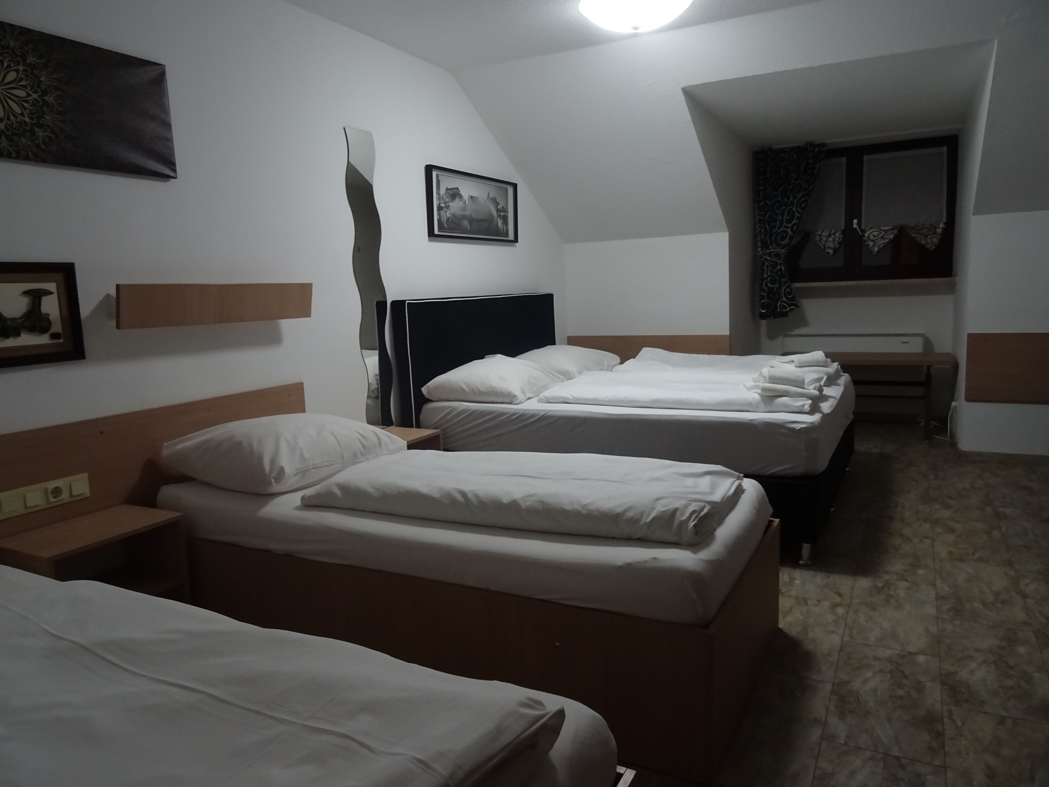 A hotel room with three single beds, a wooden headboard, a bedside table, a wall-mounted picture, and a mirror with curtains.