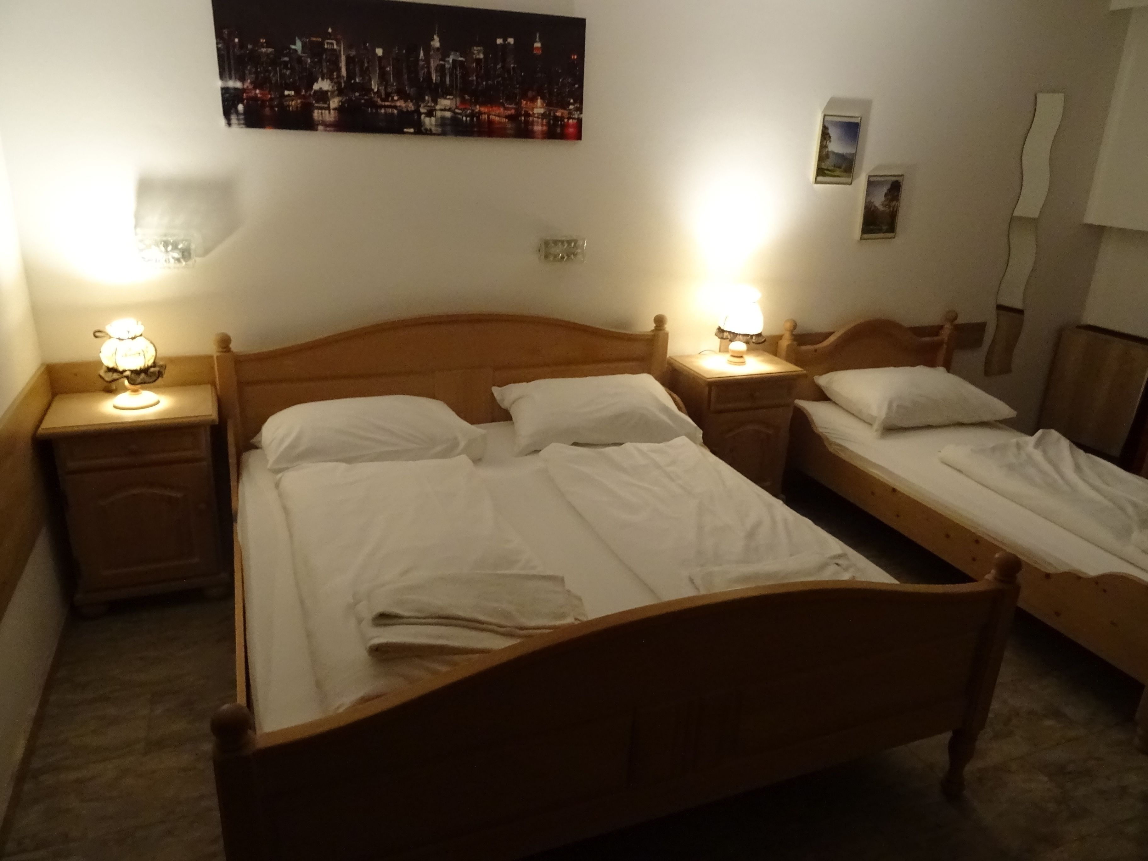 Habitación de hotel con una cama grande, dos mesitas de noche con lámparas, un mueble auxiliar y una pintura de un paisaje urbano en la pared.