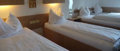 Habitación de hotel con tres camas individuales, cada una con ropa blanca y almohadas. Hay lámparas fijadas a la pared sobre cada cama.